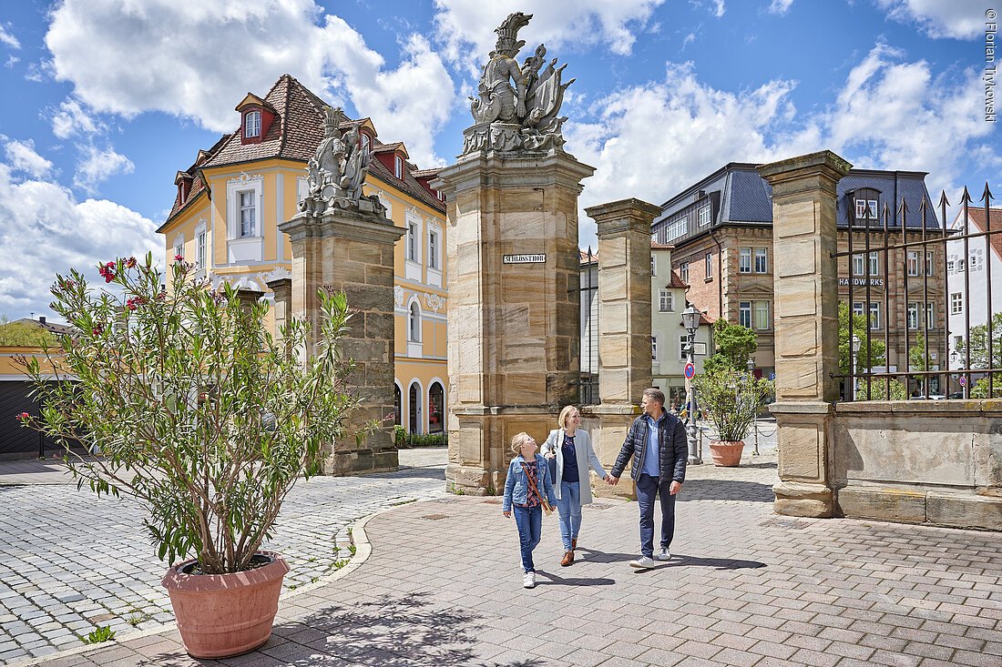 Drei Personen, wahrscheinlich eine Familie, schlendern vor einem großen, vornehm aussehenden Haus durch eine frühlingshafte Stadt