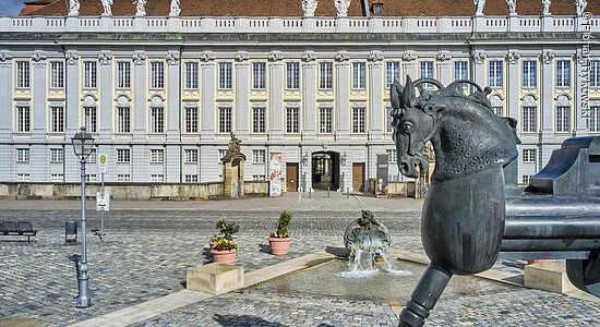 Ein großes, barockes Gebäude in bläulicher Farbe, davor eine prunkvolle Zufahrt und ein Pferd als Kunstwerk