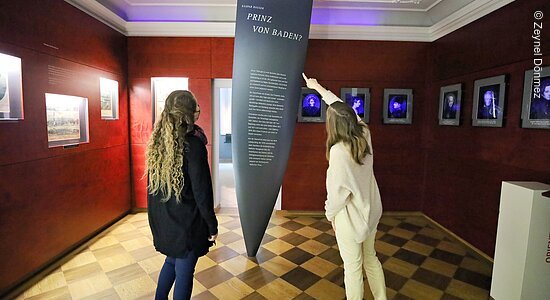 Zwei Personen deuten auf ein großes Ausstellungsstück in einem roten Museumsraum