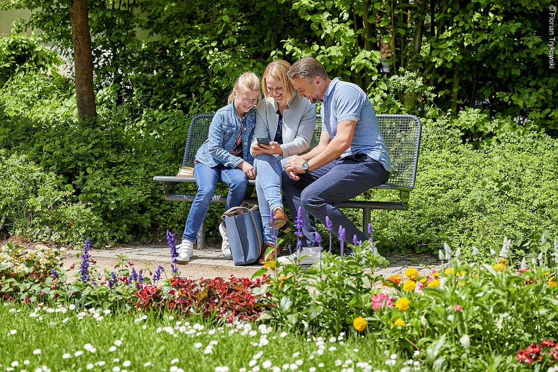 Drei Leute sitzen eng zusammen auf einer Bank in einem wunderbar grünen, blühenden Park