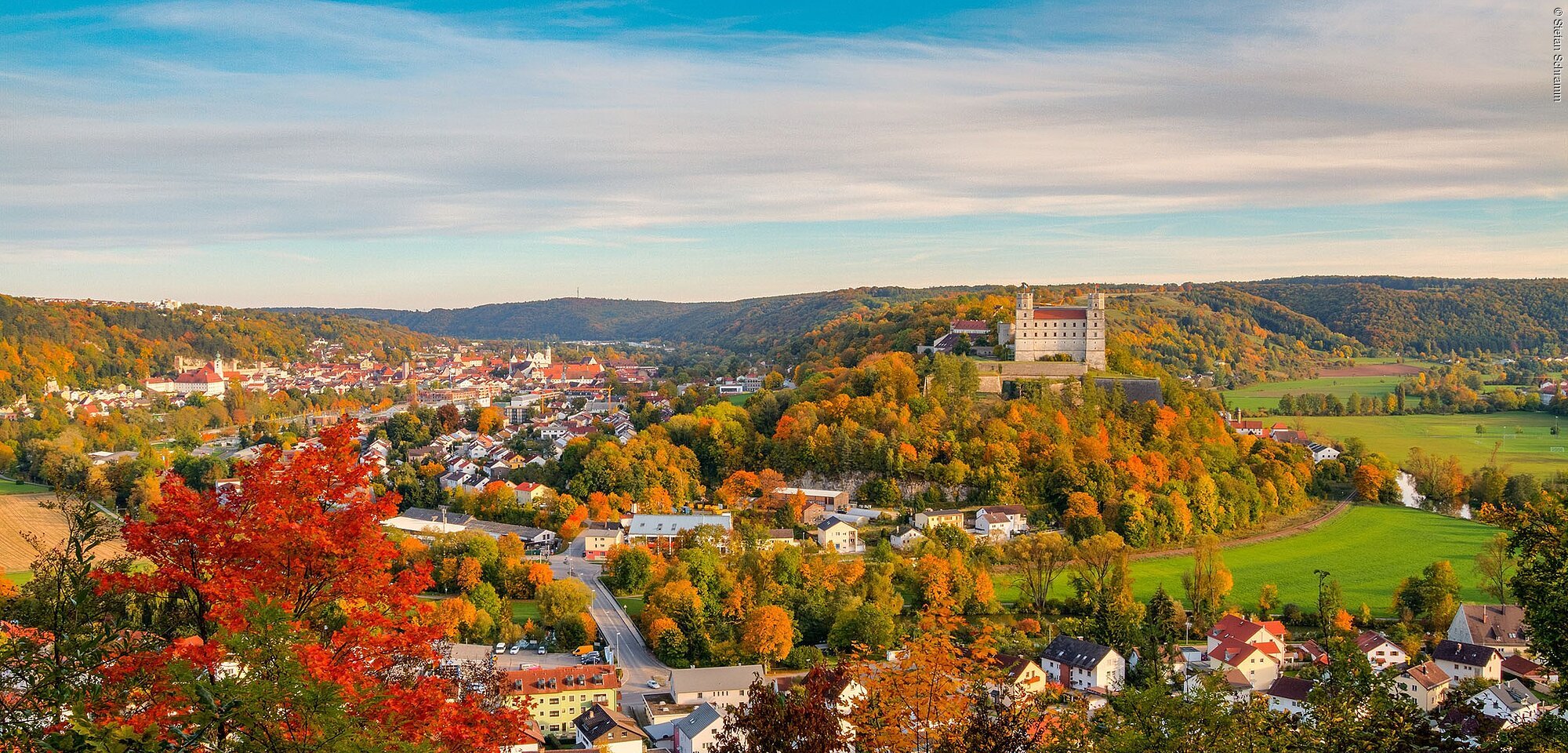 Blick über eine Stadt mit Burg, umgeben von herbstlichen Wäldern