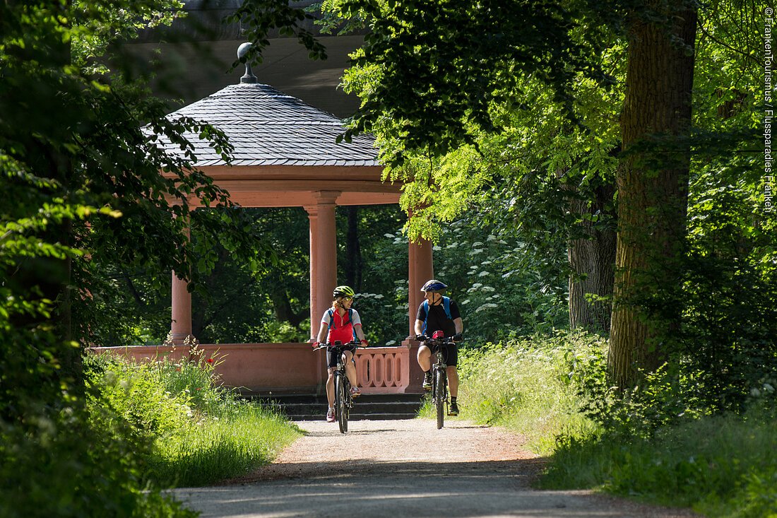 Zwei Radelnde umgeben von Grün, im Hintergrund ein Pavillon