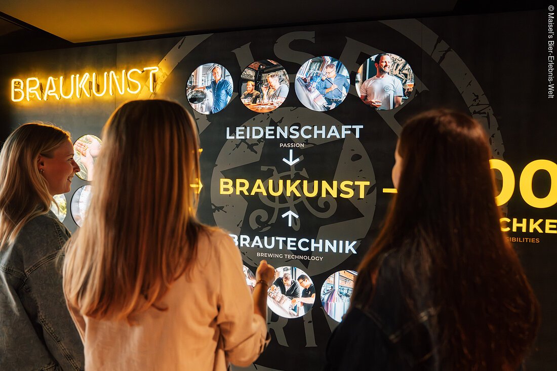Drei Personen betrachten eine beleuchtete Wand mit dem Wort "BRAUKUNST" und Bildern von Brauereien.