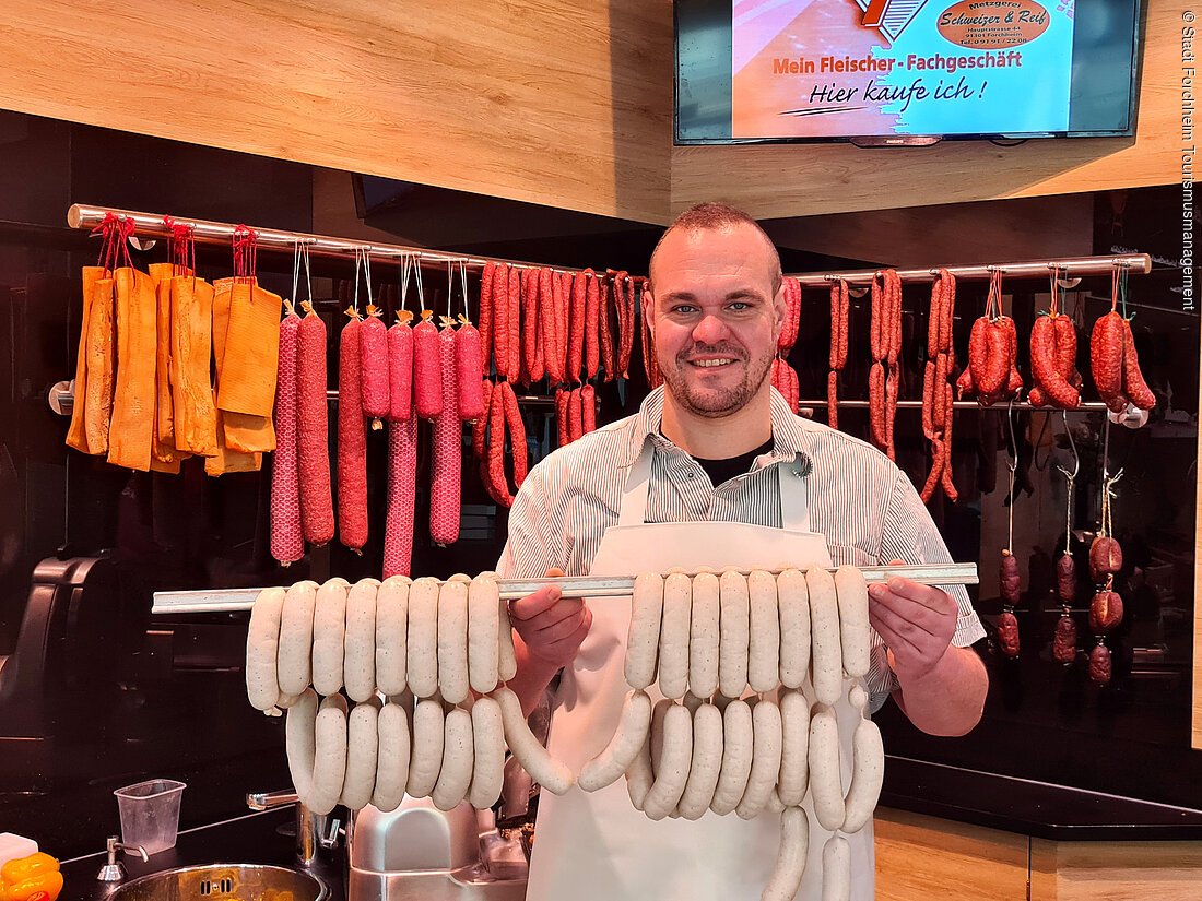 Mann mit Schürze hält zwei Stangen mit Weißürsten vor einer Theke mit hängenden Würsten.