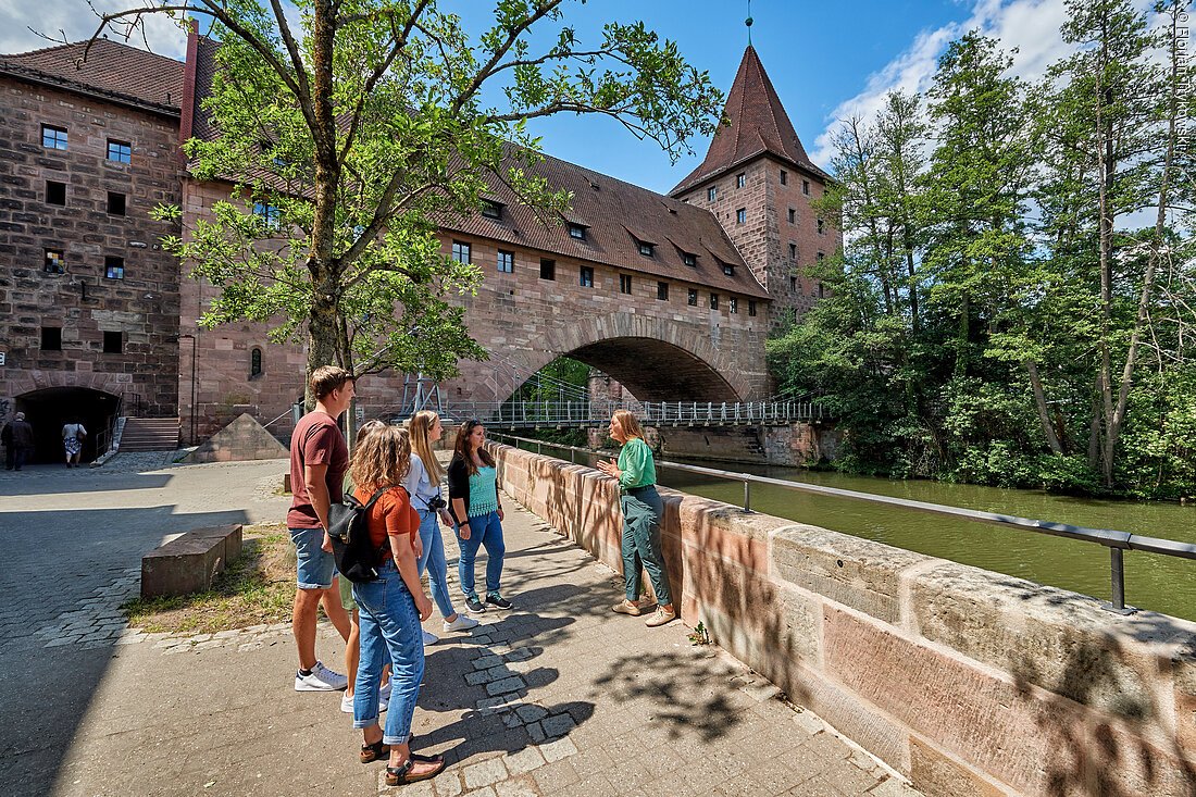 Fünf Personen stehen an einer Steinmauer an einem Fluss vor einer historischen Brücke mit Turm bei sonnigem Wetter.