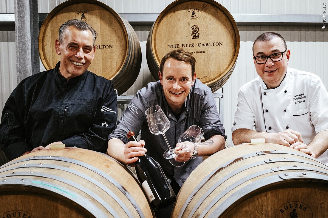 Drei Männer in Kochkleidung sitzen lächelnd hinter Weinfässern, einer hält Weinflasche und Gläser.