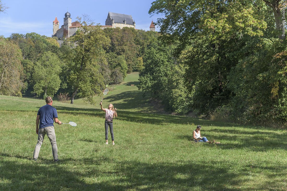 Mann und Mädchen spielen Badminton auf Wiese, Frau sitzt lesend im Gras, Burg im Hintergrund