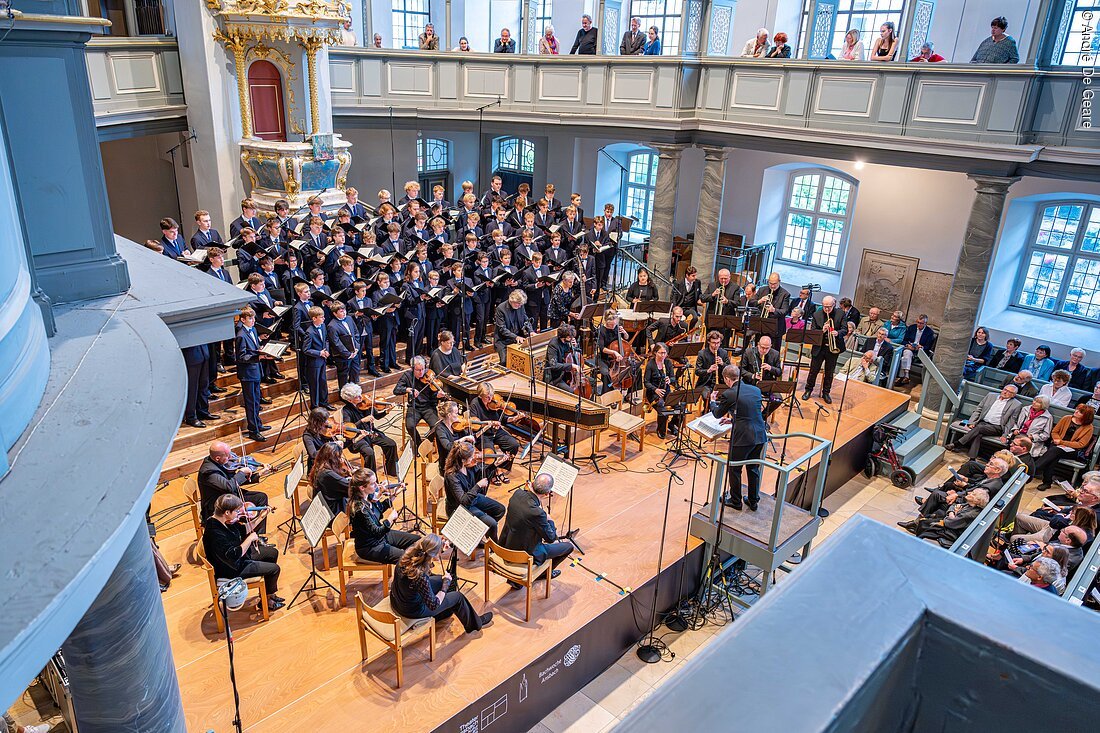 Orchester und Chor bei Konzert in einer Kirche mit Publikum auf Empore und Bänken.
