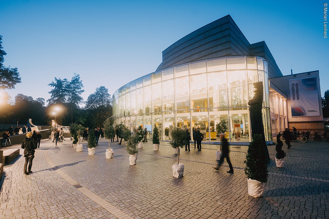 Abendaufnahme eines modernen Gebäudes mit großer Glasfassade und Menschen auf gepflastertem Vorplatz mit Bäumen in Töpfen.