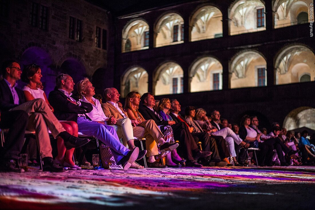 Publikum sitzt in Reihen bei Nacht vor beleuchtetem historischen Gebäude mit Arkaden und beobachtet Veranstaltung.
