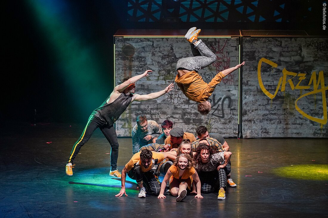 Tänzer führen eine akrobatische Formation auf einer Bühne mit Graffiti-Hintergrund aus.