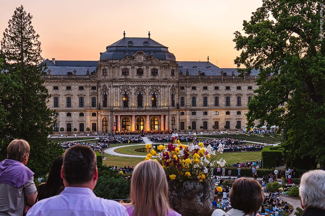 Menschen schauen bei Sonnenuntergang auf ein beleuchtetes Schloss mit Konzert im Garten davor.