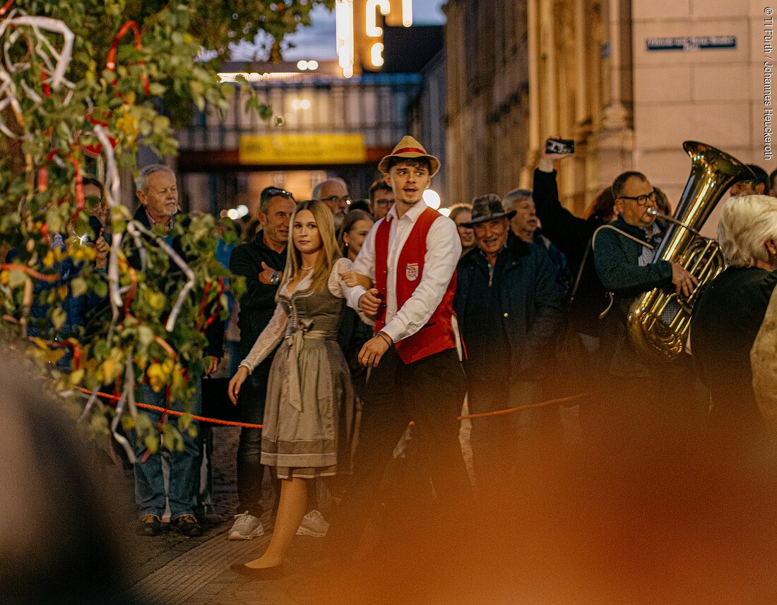 Junges Paar in traditioneller Tracht bei Abendveranstaltung mit Zuschauern und Musiker in Stadtstraße.