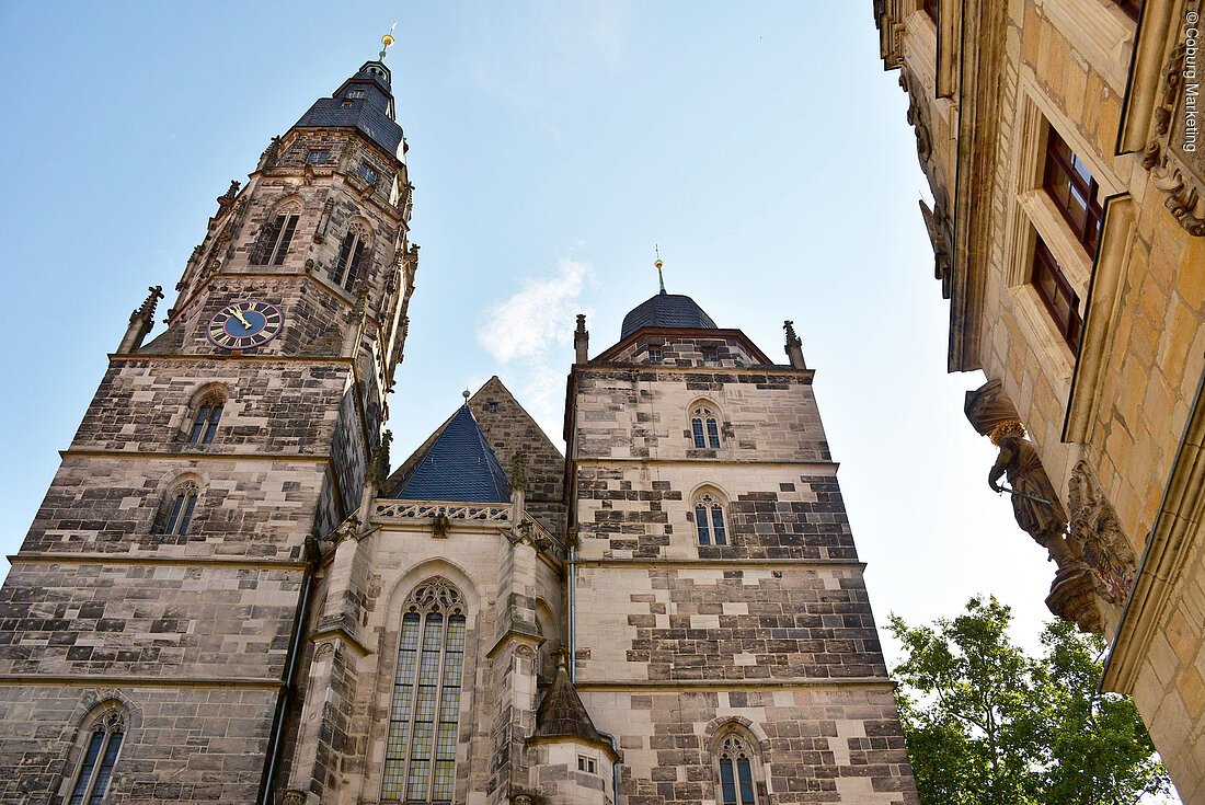 Gotische Kirche mit Uhrturm und Spitzdach vor blauem Himmel, rechts Gebäudefassade mit Statue und Baum sichtbar