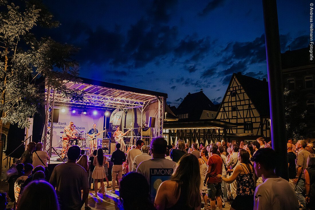 Menschenmenge vor Bühne mit Band bei Nacht in einem Ort mit Fachwerkhäusern.
